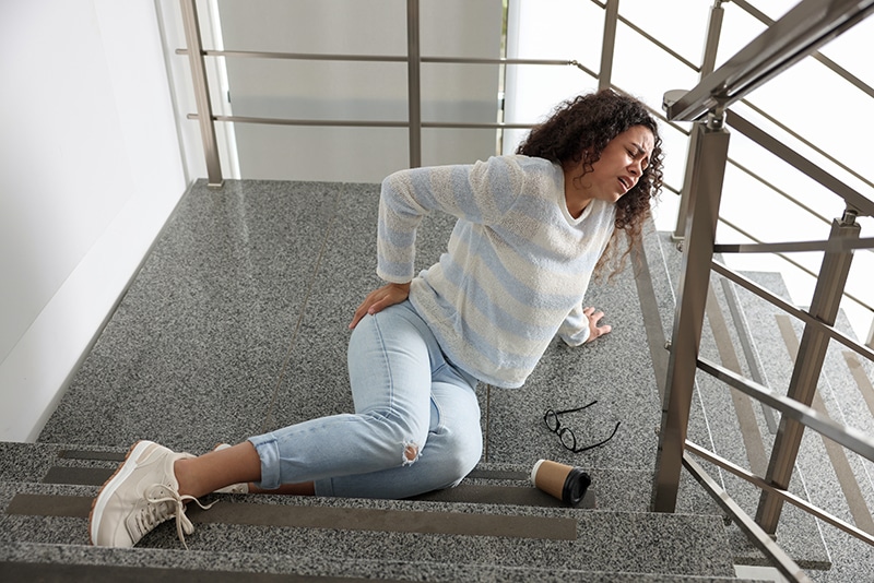 Mujer cayendo por unas escaleras en un edificio, sosteniéndose la espalda tras la caída.